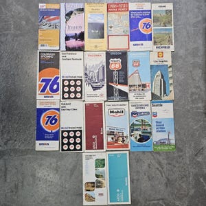 May include: A collection of twenty-two vintage paper road maps and travel guides featuring various regions, cities, and gas station brands like 76, Mobil, Phillips 66, and Chevron. The rectangular maps display colorful illustrations, text, and logos.