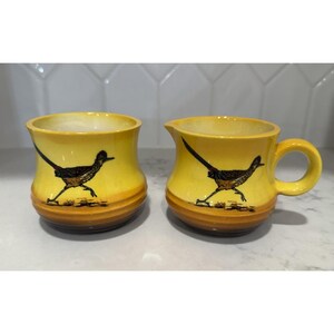 May include: Two yellow ceramic mugs featuring a roadrunner bird design. One mug has a handle. The roadrunner is depicted in black and brown, with a long tail and running pose. The mugs have a brown band at the base.