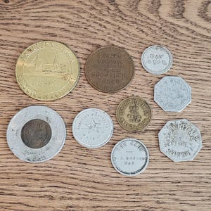 Puede incluir: Una colección de fichas y monedas antiguas, que incluye una ficha Sandusky Sesquicentennial dorada con una ilustración de barco y una ficha Golden Virginia Good Luck. Otras fichas presentan texto y diseños de estrellas.