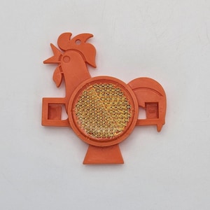 Op de afbeelding: Een oranje plastic reflector in de vorm van een haan, met een cirkelvormig, goudkleurig reflecterend midden. De haan heeft een gedetailleerde kam, snavel en staart. De reflector is ontworpen om aan een fiets of ander object te worden bevestigd.