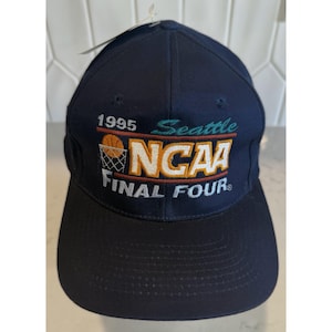 Puede incluir: Una gorra de béisbol azul marino con un logotipo bordado de 1995 Seattle NCAA Final Four en la parte delantera. El diseño incluye un gráfico de baloncesto y aro, con el texto 1995, Seattle, NCAA y FINAL FOUR bordado en blanco, oro y verde azulado.