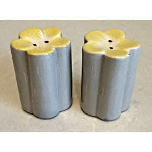 Puede incluir: Un par de saleros y pimenteros de cerámica. Cada salero y pimentero tiene un cuerpo cilíndrico gris y una parte superior en forma de flor amarilla. Las partes superiores de las flores tienen dos agujeros para dispensar el contenido.