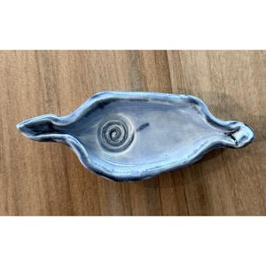 May include: A handmade, light blue ceramic dish with a spiral design in the center. The dish has a unique, elongated shape with curved edges and a glossy finish. The ceramic dish is a decorative item.