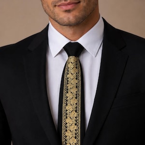 May include: A black necktie featuring a central vertical panel of intricate gold floral embroidery. The tie is worn with a crisp white collared shirt and a solid black suit jacket, creating a formal appearance with a distinct metallic gold accent.