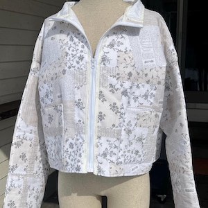 May include: A white quilted jacket with a floral and newspaper print. It has a full-length white zip, two front pockets, and a stand-up collar. The long-sleeved jacket is displayed on a mannequin, showcasing its design.