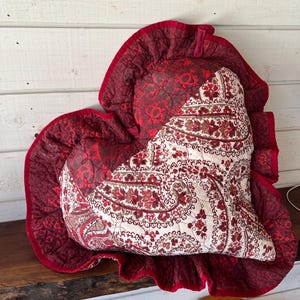 May include: A heart-shaped quilted pillow featuring a deep red fabric with a dark floral pattern on one side and a cream-colored fabric with a detailed red and brown paisley print on the other, finished with a ruffled red fabric border trim.