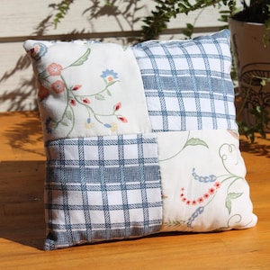 May include: A square throw pillow featuring a patchwork design of four alternating fabric panels. Two panels display a white background with a blue plaid pattern, while the other two panels feature a white background with floral vines.