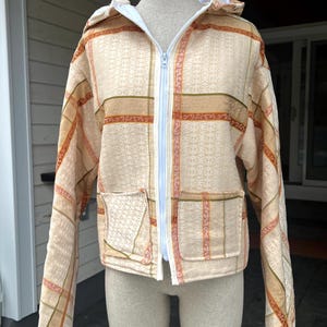 May include: Beige hooded jacket with a plaid pattern in shades of orange, pink, and green. The jacket has a white zipper and two front pockets. The jacket is displayed on a mannequin.