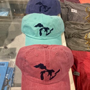Könnte beinhalten: Eine Auswahl an Baseballkappen in verschiedenen Farben, darunter Rot, Blau und Türkis. Jede Kappe zeigt ein dunkelblaues, gesticktes Design eines Seeumrisses. Die Kappen sind in einer Reihe angeordnet, um die verschiedenen Farboptionen zu präsentieren.