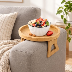 May include: A round bamboo sofa arm tray with foldable legs holding a white ribbed ceramic bowl filled with oatmeal, fresh strawberries, blueberries, and cashews. The tray features a light wood grain finish and a stable design for armrests.