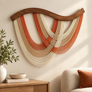 May include: A wall hanging featuring a curved dark wood frame with multiple layers of draped rope in cream, tan, and burnt orange colors. The ropes are arranged in overlapping, cascading U-shaped patterns suspended from the wooden structure.