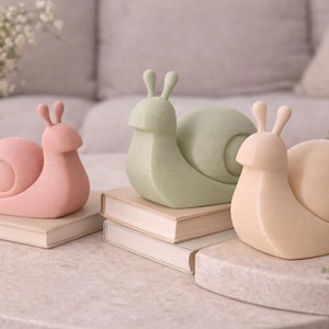 May include: Three decorative snail figurines in pastel colors: pink, green, and beige. Each snail sits atop a book on a light-colored table. The snails have a smooth, rounded design, with visible shell details and antennae. Home decor.