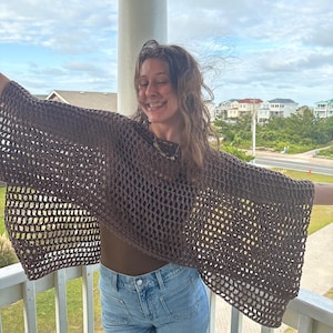 Puede incluir: Un top de crochet marrón de tejido abierto con mangas anchas. El top está hecho de hilo marrón oscuro y tiene un diseño suelto y aireado. El top se usa con un top marrón y jeans azules.