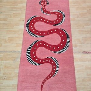 Puede incluir: Una alfombra rectangular de color rosa empolvado con un gran motivo de serpiente sinuosa. La serpiente es de color rojo oscuro con lunares blancos y bordes a rayas negras y blancas. La alfombra tiene un borde con flecos en los extremos.
