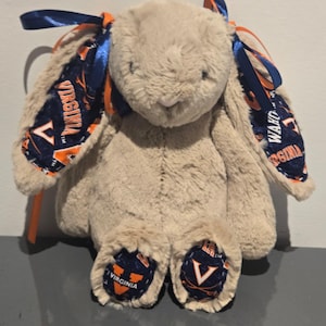 May include: A plush bunny rabbit in a light brown color. The bunny's ears and feet feature a navy blue and orange patterned fabric with the word "VIRGINIA" printed on it. The bunny has blue ribbons tied to its ears.
