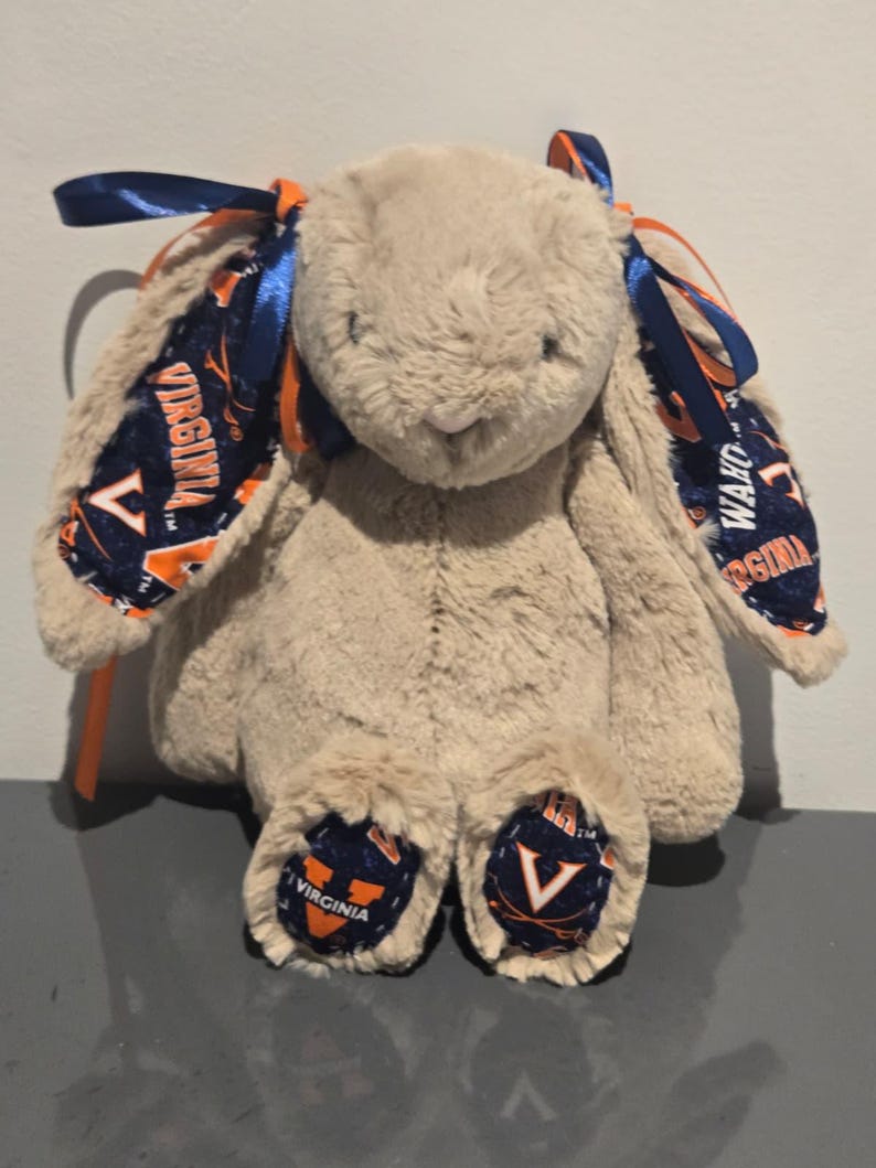May include: A plush bunny rabbit in a light brown color. The bunny's ears and feet feature a navy blue and orange patterned fabric with the word "VIRGINIA" printed on it. The bunny has blue ribbons tied to its ears.