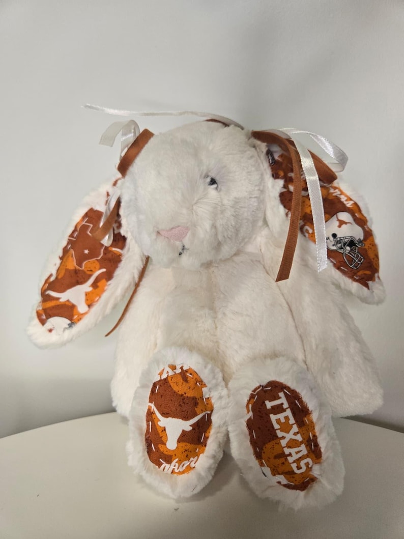 May include: A plush white bunny with brown and orange patterned ears and feet. The ears and feet feature a repeating pattern of the Texas Longhorns logo and the word "TEXAS". The bunny has brown ribbon accents and a pink nose.