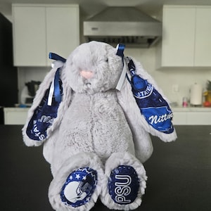 May include: A plush gray bunny with blue accents. The bunny has long ears with blue and white patterned fabric, blue bows, and patches on its feet with the letters "PSU" and a football helmet design. The bunny has a pink nose.