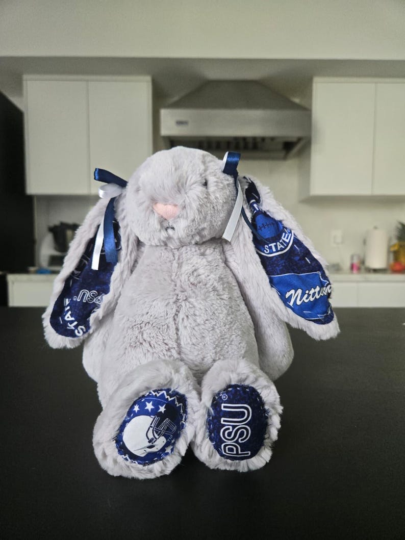 May include: A plush gray bunny with blue accents. The bunny has long ears with blue and white patterned fabric, blue bows, and patches on its feet with the letters "PSU" and a football helmet design. The bunny has a pink nose.
