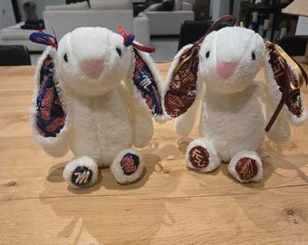 May include: Two white plush bunny rabbits with pink noses and patterned ears. One bunny has blue and red accents, the other has maroon and gold accents. The bunnies have matching accents on their feet.