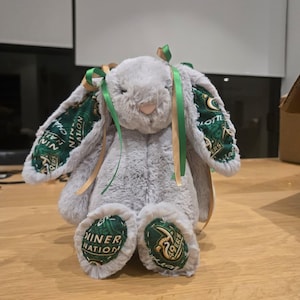May include: A plush gray bunny with green and gold accents. The bunny's ears and feet feature a green fabric with white text and gold accents. Green and gold ribbons are tied to the ears. The bunny is sitting on a wooden surface.