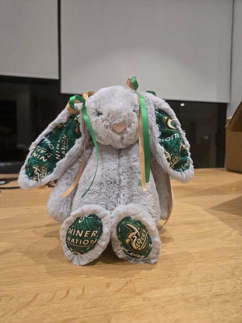 May include: A plush gray bunny with green and gold accents. The bunny's ears and feet feature a green fabric with white text and gold accents. Green and gold ribbons are tied to the ears. The bunny is sitting on a wooden surface.