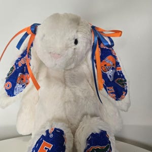 May include: A white plush bunny with blue and orange accents. The bunny's ears and feet feature a blue fabric with orange and white graphics. Blue and orange ribbons are tied to the ears. The bunny is sitting on a white surface.