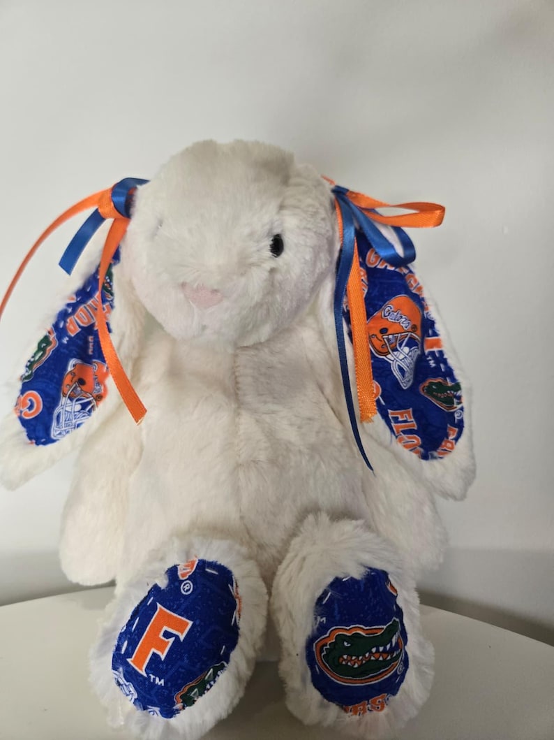 May include: A white plush bunny with blue and orange accents. The bunny's ears and feet feature a blue fabric with orange and white graphics. Blue and orange ribbons are tied to the ears. The bunny is sitting on a white surface.