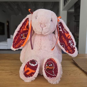 May include: A plush pink bunny with long ears and a soft, fluffy texture. The inner ears and foot pads feature a maroon and orange pattern with the text "VIRGINIA TECH HOKIES". Orange and maroon ribbons adorn the ears.