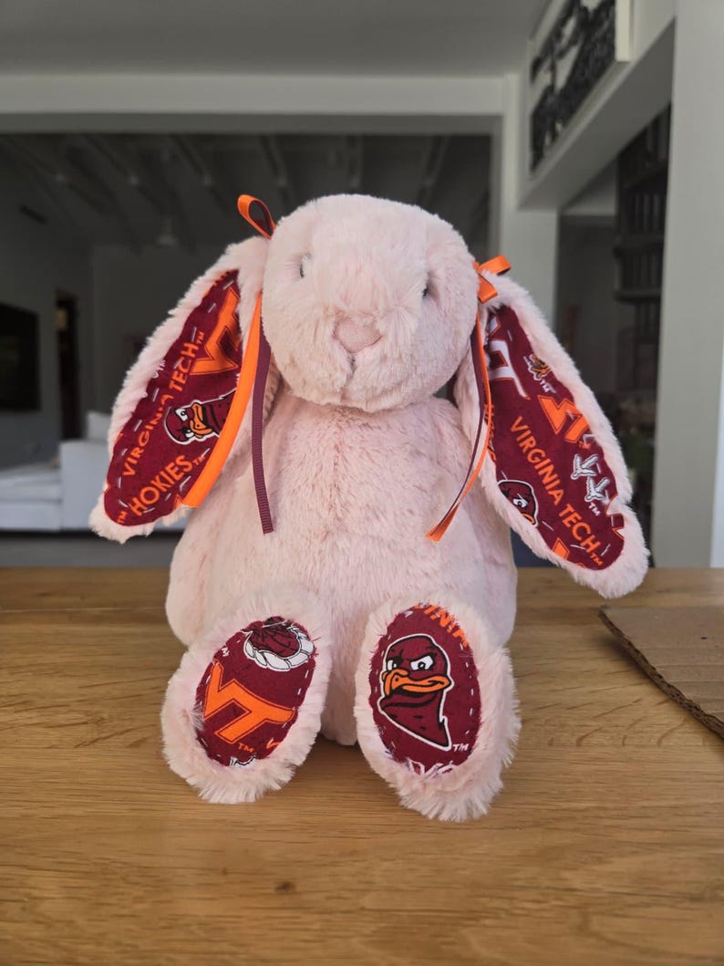 May include: A plush pink bunny with long ears and a soft, fluffy texture. The inner ears and foot pads feature a maroon and orange pattern with the text "VIRGINIA TECH HOKIES". Orange and maroon ribbons adorn the ears.