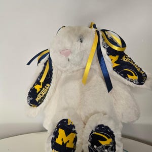 May include: A white plush bunny with blue and yellow accents. The bunny's ears and feet feature a navy blue fabric with the word "MICHIGAN" and a yellow "M" logo. Yellow and blue ribbons are tied to the ears.