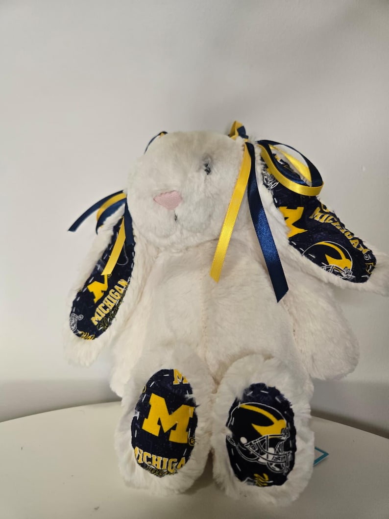 May include: A white plush bunny with blue and yellow accents. The bunny's ears and feet feature a navy blue fabric with the word "MICHIGAN" and a yellow "M" logo. Yellow and blue ribbons are tied to the ears.