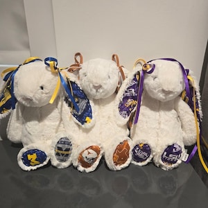 May include: Three white plush bunny rabbits with team logos on their feet and ears. Each bunny has a different team logo, including the University of Michigan, the University of Texas, and LSU Tigers. The bunnies have colorful ribbons.