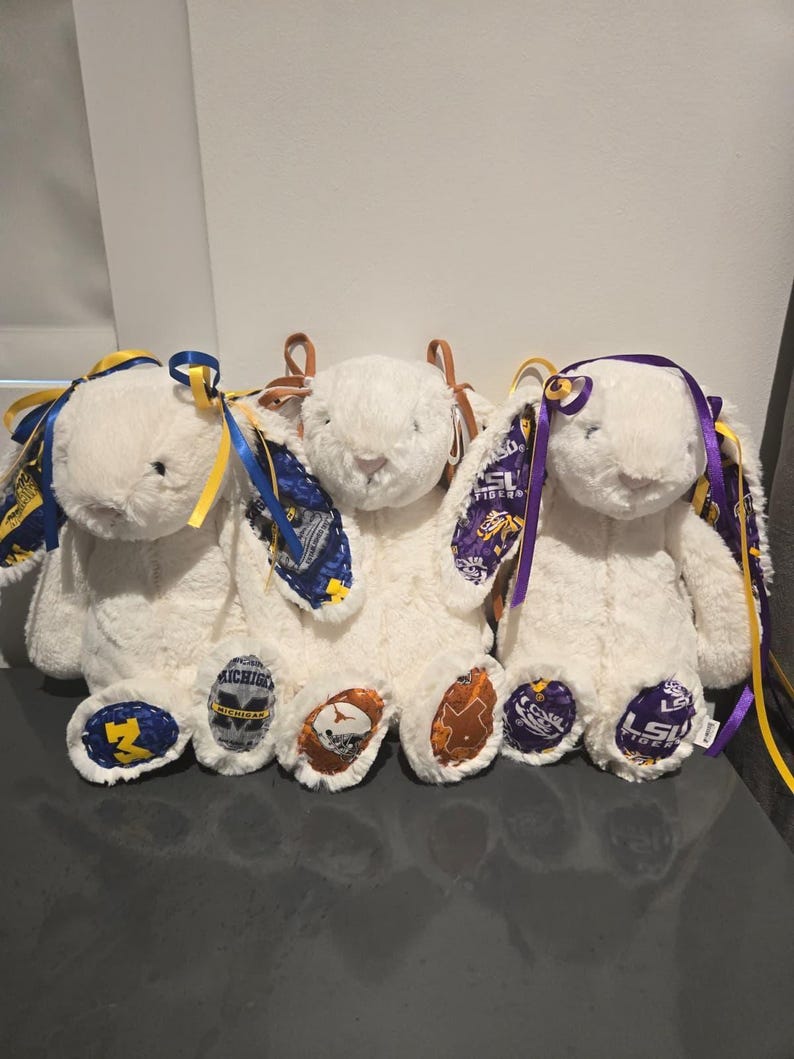 May include: Three white plush bunny rabbits with team logos on their feet and ears. Each bunny has a different team logo, including the University of Michigan, the University of Texas, and LSU Tigers. The bunnies have colorful ribbons.