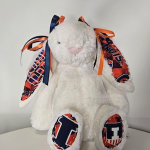 May include: A plush white bunny with orange and navy blue accents. The bunny's ears are adorned with patterned fabric and ribbons in matching colors. The feet feature a navy blue "I" logo on a red background. A soft, cuddly toy.