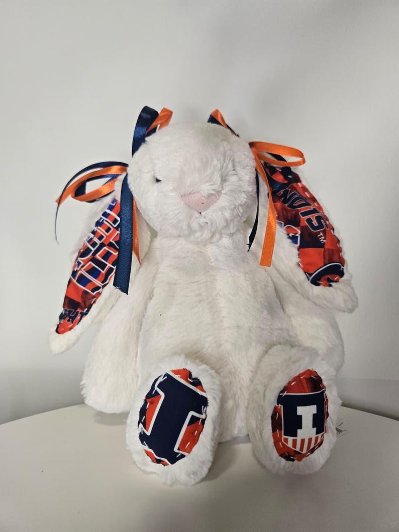 May include: A plush white bunny with orange and navy blue accents. The bunny's ears are adorned with patterned fabric and ribbons in matching colors. The feet feature a navy blue "I" logo on a red background. A soft, cuddly toy.