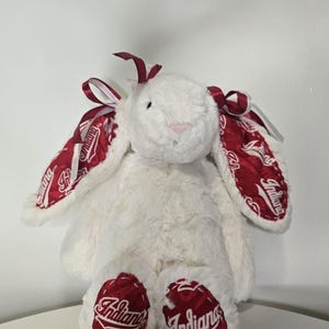 May include: A plush white bunny with red accents. The bunny's ears and feet feature a red and white design with the word "Indiana". The bunny has a pink nose and red ribbon accents.