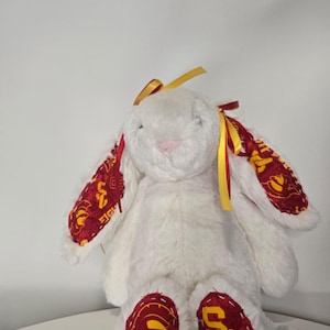 May include: A white plush bunny with long ears and a pink nose. The bunny's ears and feet feature a maroon and gold patterned fabric. Yellow and red ribbons adorn the ears. A cute stuffed animal.