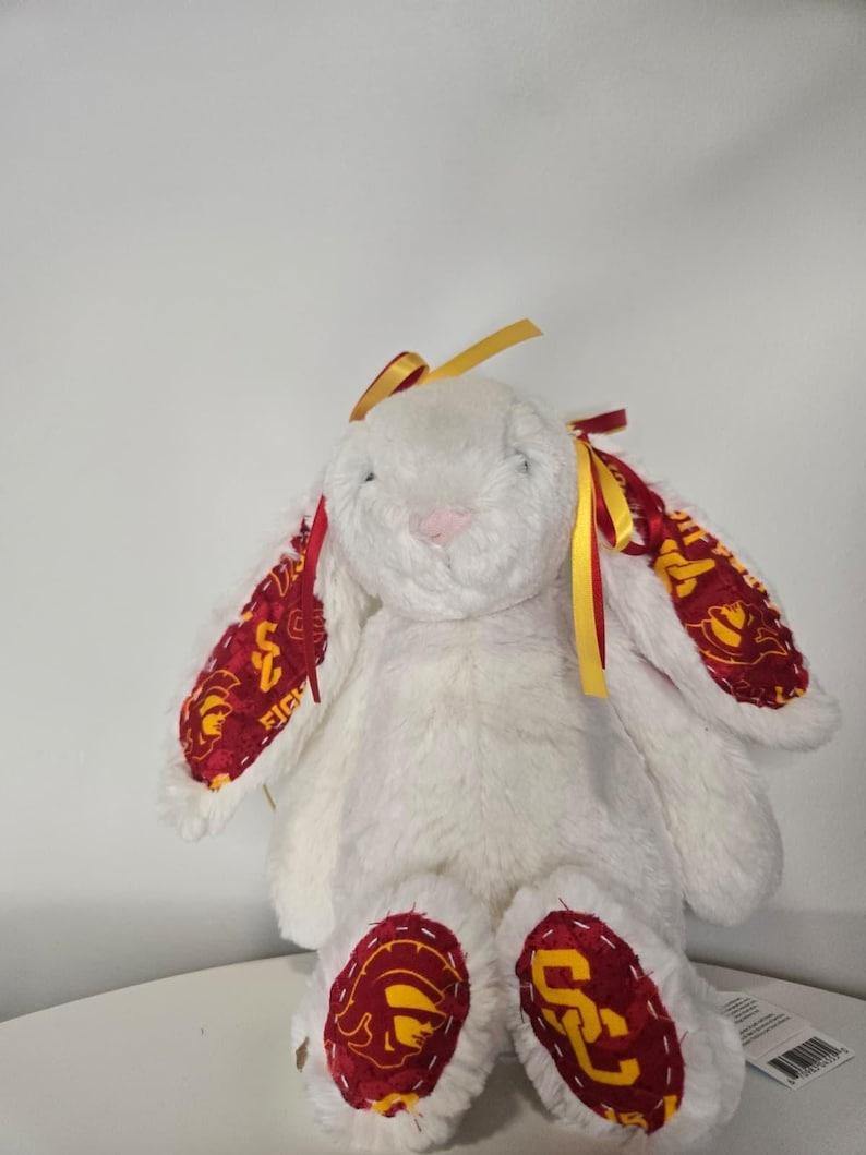 May include: A white plush bunny with long ears and a pink nose. The bunny's ears and feet feature a maroon and gold patterned fabric. Yellow and red ribbons adorn the ears. A cute stuffed animal.
