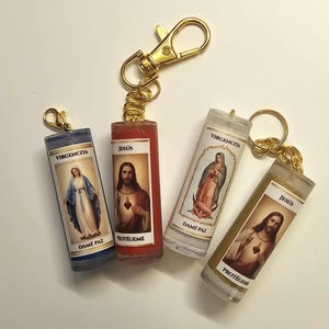 May include: Four cylindrical keychains featuring religious imagery. Two depict the Virgin Mary with the text VIRGENCITA and DAME PAZ, while two depict Jesus with the text JESÚS and PROTÉGEME. Each keychain has a gold-toned metal clasp and chain.