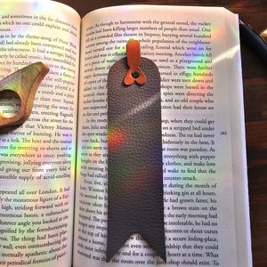 May include: A dark gray leather bookmark with a unique shape, featuring a small orange accent and a leather loop. The bookmark is resting on the pages of a book, adding a touch of elegance to reading.