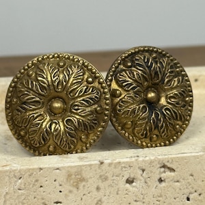 May include: Two antique brass cabinet knobs. Each knob features a detailed floral design with a central button and a beaded border. The knobs are round and have a warm, aged appearance, suggesting vintage or retro style.