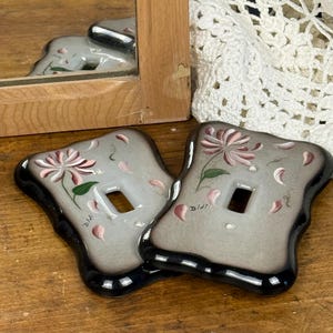 Peut inclure: Deux plaques d'interrupteur décoratives avec un motif floral. Les plaques sont grises avec une bordure noire et une fleur peinte en rose et blanc. Le mot "Didi" est écrit sur chaque plaque.