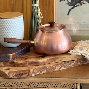 May include: A round copper pot with a polished metallic finish, featuring a long wooden handle and a matching lid with a small wooden knob. The pot has a brass-coloured attachment connecting the handle to the main body, showcasing a smooth, curved design.