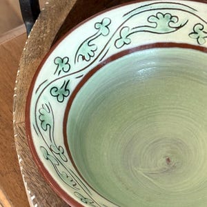 Peut inclure: Un bol en céramique avec un intérieur vert clair et un bord crème. Le bord est orné d'une bande marron et de motifs floraux verts. Le bol est posé sur un plateau en bois avec une poignée noire.