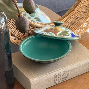 May include: A teal ceramic bowl with a floral-decorated lid. The lid features a design in yellow, brown, and blue. The bowl is open, revealing its interior. The bowl and lid are resting on a book with the words "Savoir Vivre" printed on the spine.