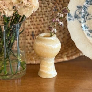 Op de afbeelding: Een kleine vaas van gepolijste steen met een rond lichaam en een uitlopende voet, voorzien van horizontale banden in crème, beige en wit. De vaas heeft een gladde, glanzende afwerking en een smalle opening aan de bovenkant met een bloemstengel.