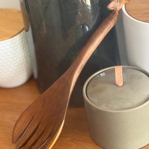 May include: A wooden salad server with a giraffe head design on the handle. The server is made of a light brown wood and has a fork-like end. The giraffe head is carved in detail. The server is on a wooden surface.