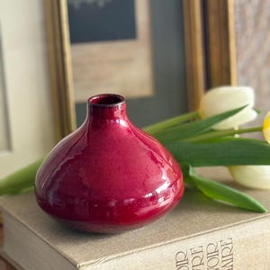 May include: A glossy, ruby-red vase with a rounded body and a narrow neck. The vase sits atop a book with gold lettering, with a few white and yellow tulips in the background. The vase is a decorative item.