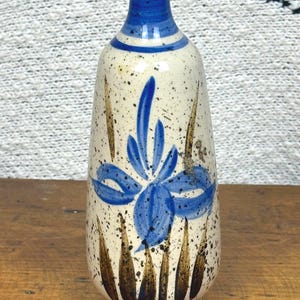 Könnte beinhalten: Eine Keramikvase mit blauem Blumendesign. Die Vase hat einen gesprenkelten cremefarbenen Körper mit braunen und blauen Akzenten. Der Hals und das Blumendesign sind blau. Die Vase steht auf einer Holzoberfläche.